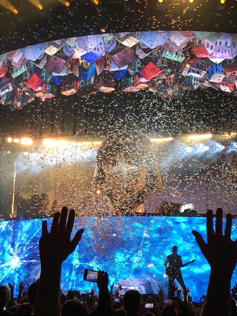 AJR performing in a stadium. Confetti is raining down. A blow-up head of one of the performers is between to very large movable screens. The bottom screen is a water-like image. The top screen is a large illustration of dense single-family detached houses upside down.