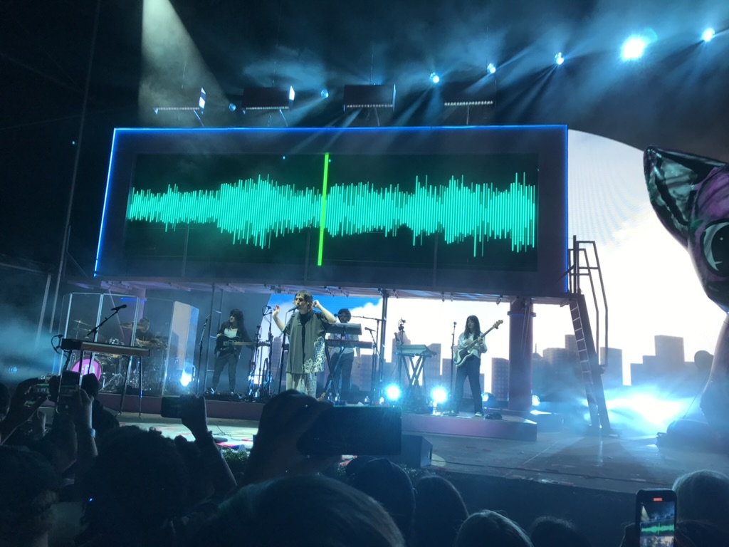 Porter Robinson on stage with an LED billboard behind him, like one you'd see on a freeway but its legs are short and there is a ladder to get up. Behind Robinson are multiple supporting musicians.