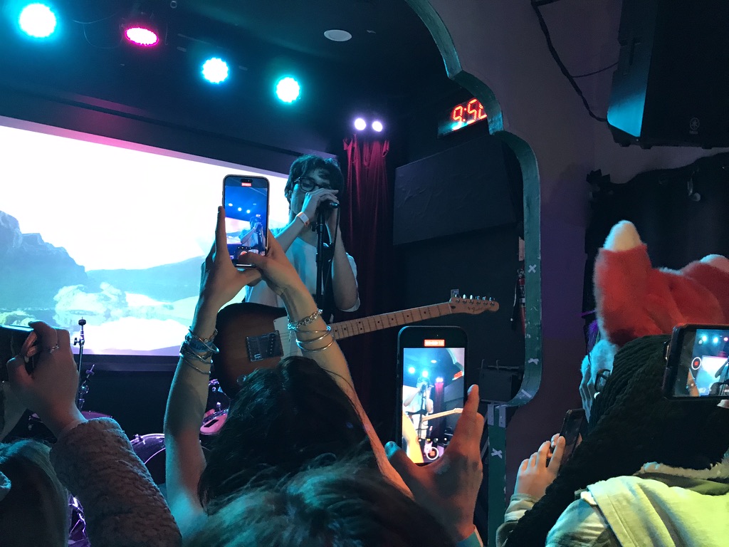 A man is singing into a microphone. There are a lot of people with phones up recording him. There is a furry with their head on, off to the side.