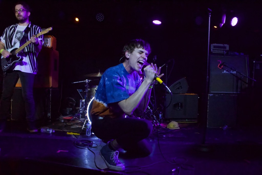Lincoln Kreifels yells into a microphone while crouching on stage. Lucas Hughes is standing back and to the left of him playing an electric guitar.
